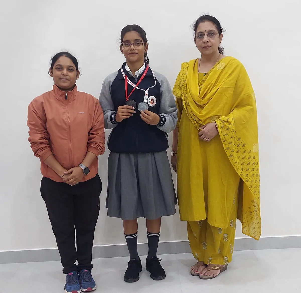 Three women standing against a white wall, with a schoolgirl in the middle holding two medals on a lanyard