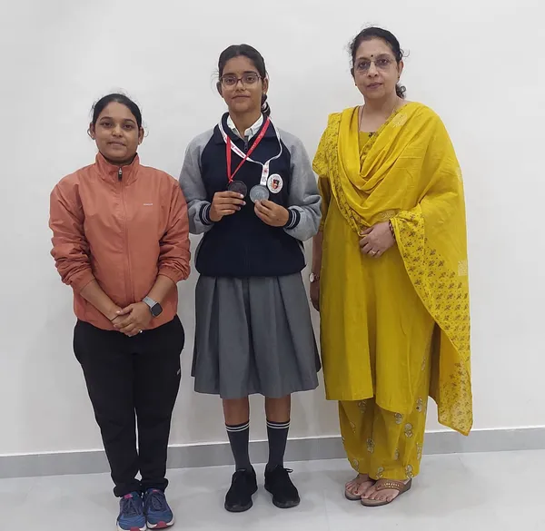 Three women standing against a white wall, with a schoolgirl in the middle holding two medals on a lanyard