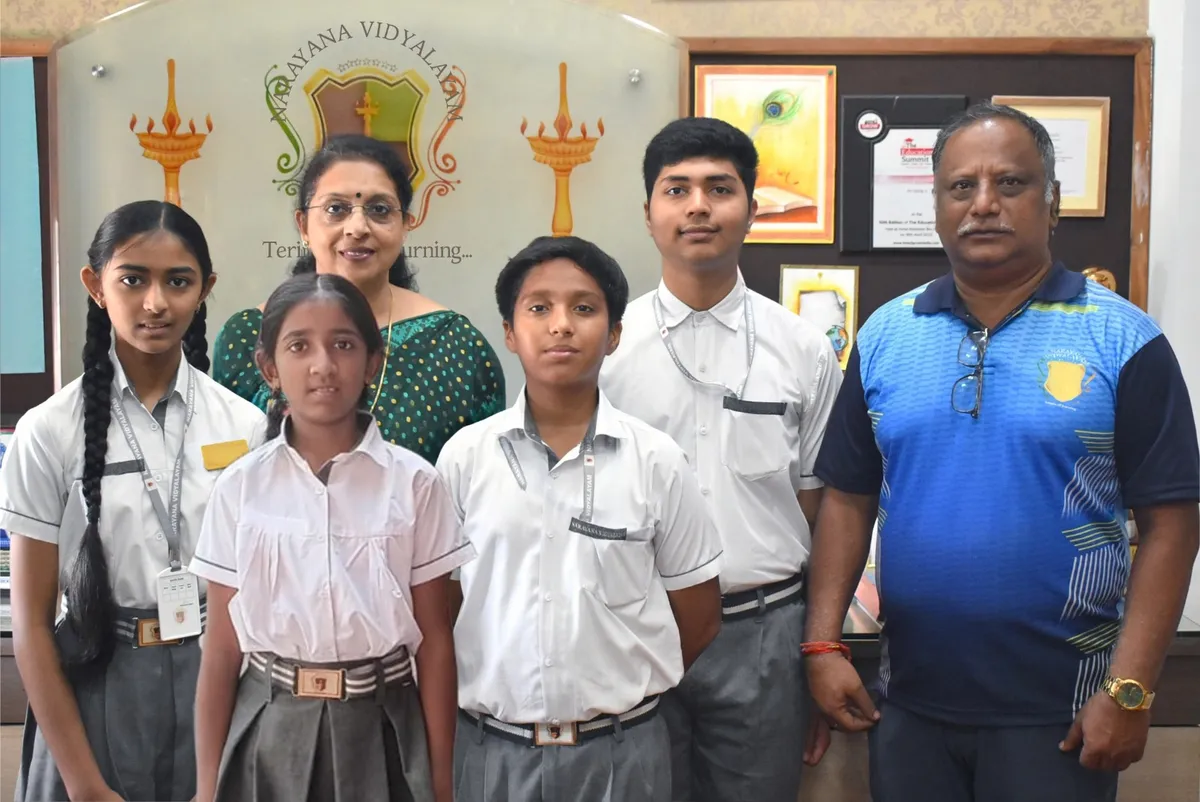Group photo of four students in school uniforms posing indoors with a woman and a man in front of a school sign and framed pictures. - Narayana Vidyalayam