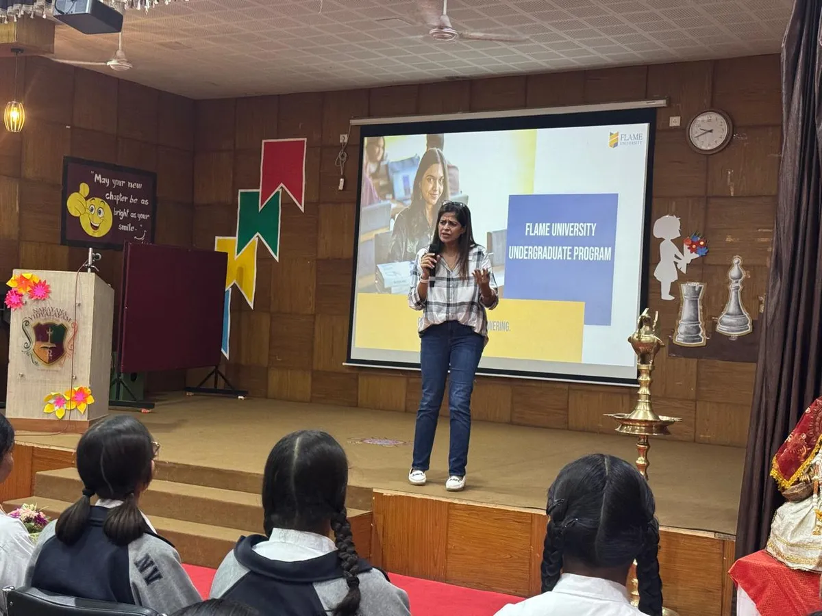 A woman with a microphone presents on a school stage in front of a projected slide while students watch from the foreground at Narayana Vidyalayam.