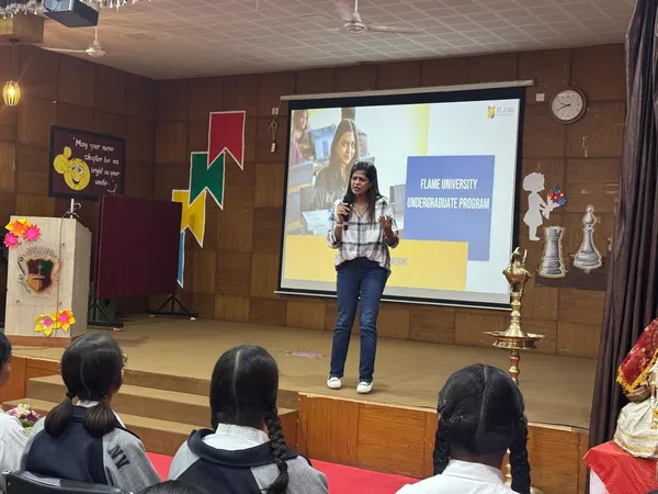 A woman with a microphone presents on a school stage in front of a projected slide while students watch from the foreground at Narayana Vidyalayam.