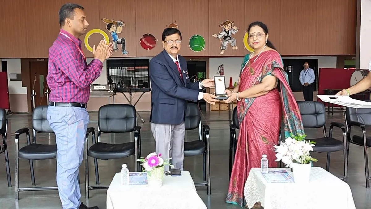 A man in a blazer hands a framed memento to a woman in a pink saree while another man applauds, inside a hall with chairs and flower-decorated tables.