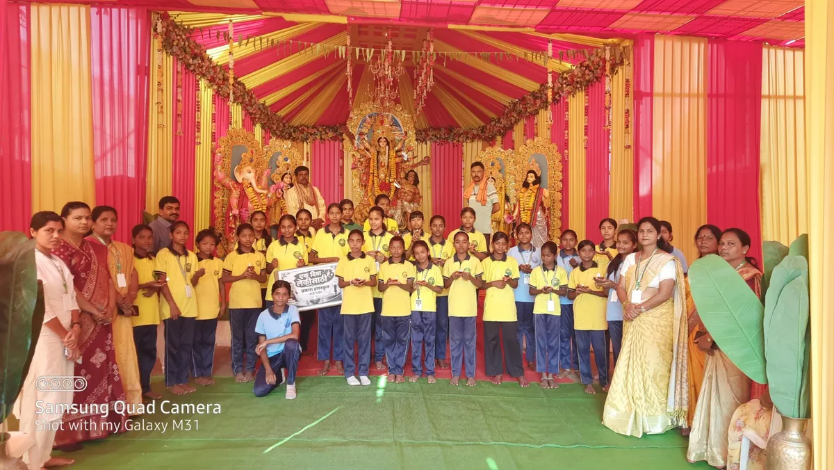 group photo of many children in yellow shirts and adults standing on a stage decorated with red and yellow drapes and religious statues, one person holding a small sign - Prakash School