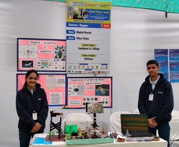 Two students in dark hoodies stand beside a science exhibition stall with a banner, posters and a small model on the table, Sandipani (Hazaripahad)