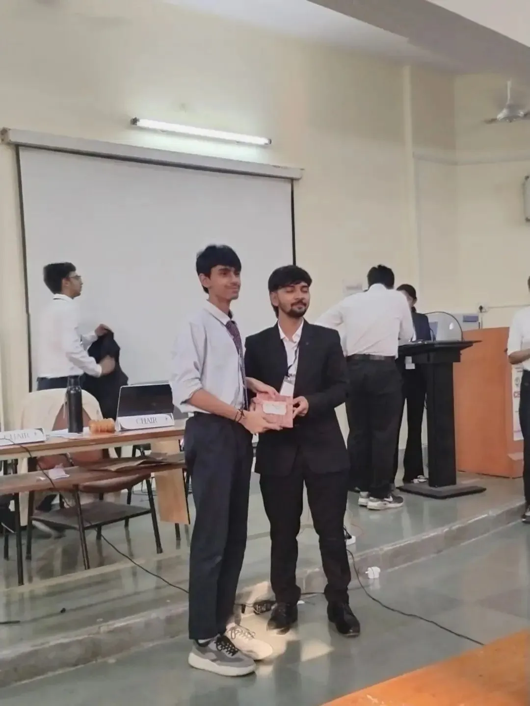 Two young men stand on a stage in a hall, one handing a small boxed item to the other, with people and a podium in the background. - Somalwar Nikalas