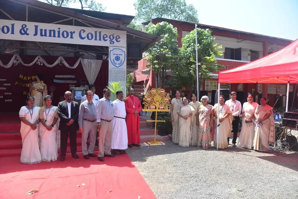 Outdoor group photo of about 17 men and women standing on a red carpet beside a decorative tree stand, in front of a school and junior college building with a stage and red tent. - St. Ursula