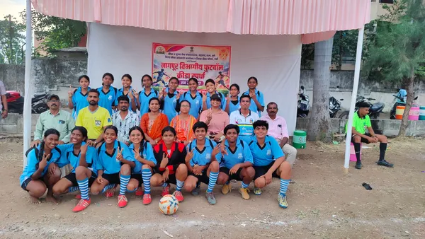 Group photo of girls in blue jerseys and several adults posing in front of a tent with a banner, with a football in the foreground. - St. Ursula