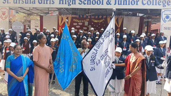 Outdoor school investiture ceremony at St. Ursula with students in uniform lined up, and teachers and a man holding blue and white house flags in the foreground.