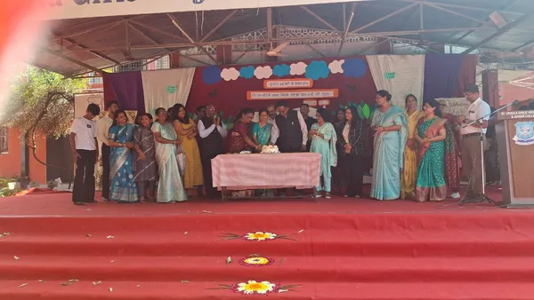 Group of women and a few men standing on a decorated stage around a table with a cake, with a banner behind them and a podium with microphone on the right. - St. Ursula