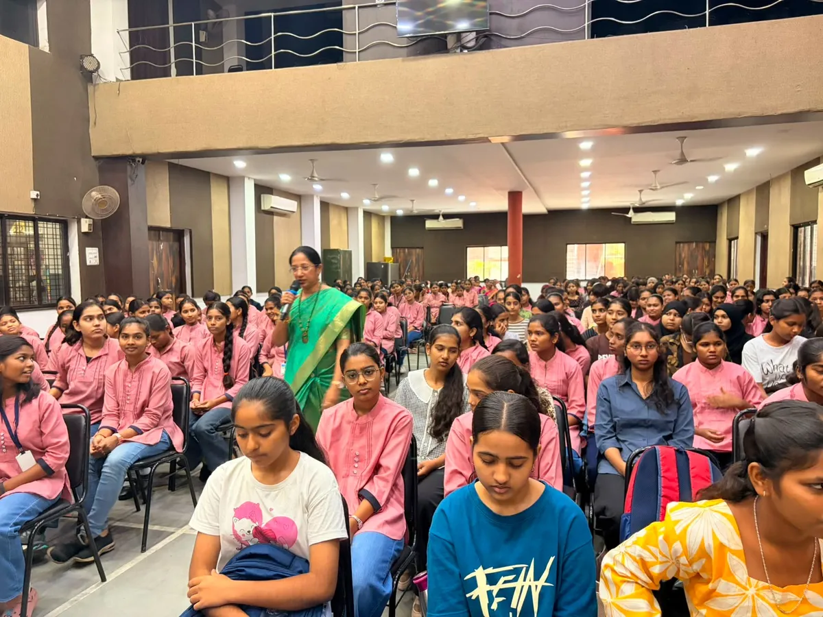 A woman speaks at a microphone in a hall while seated girls in pink uniforms listen; St. Ursula Induction Programme.
