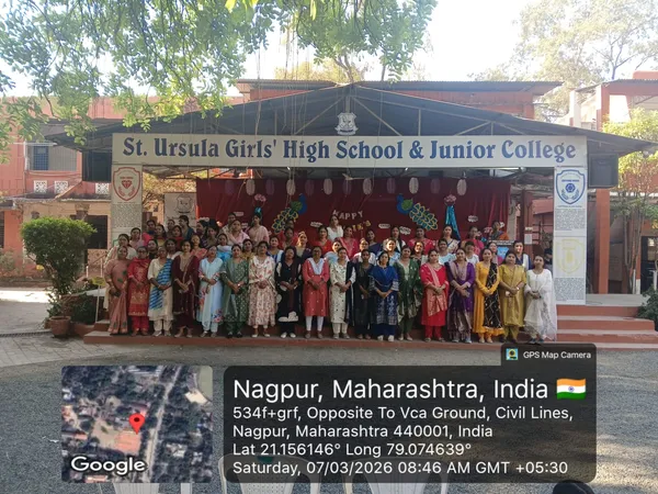 Large group photo of women in colourful traditional dress standing in rows in front of a school stage with a name board, with a GPS map overlay at the bottom. - St. Ursula