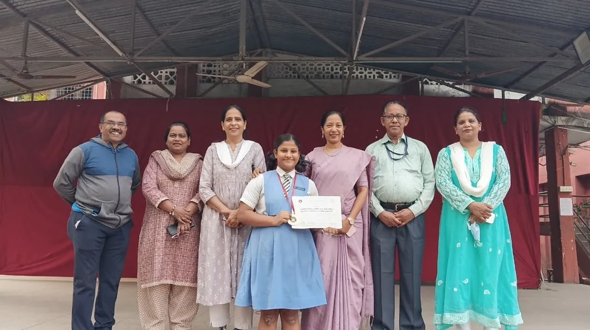 Girl in school uniform with medal and certificate standing with six adults in front of a red curtain.