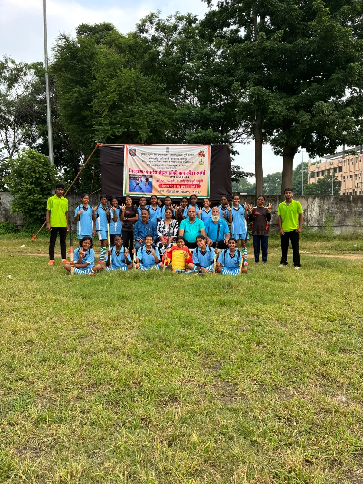 Group photo of girls in blue sports uniforms with hockey sticks and adults posed on a grass field in front of a banner and trees at St. Ursula
