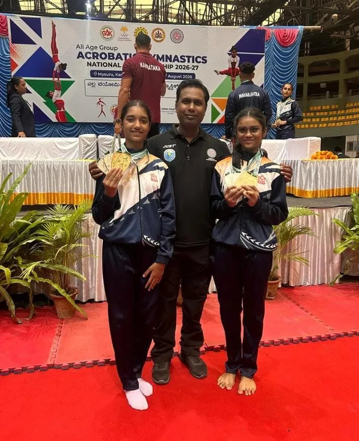 Two girls in matching tracksuits holding medals pose with a man in black between them in front of a gymnastics championship banner on a red mat at St. Ursula.