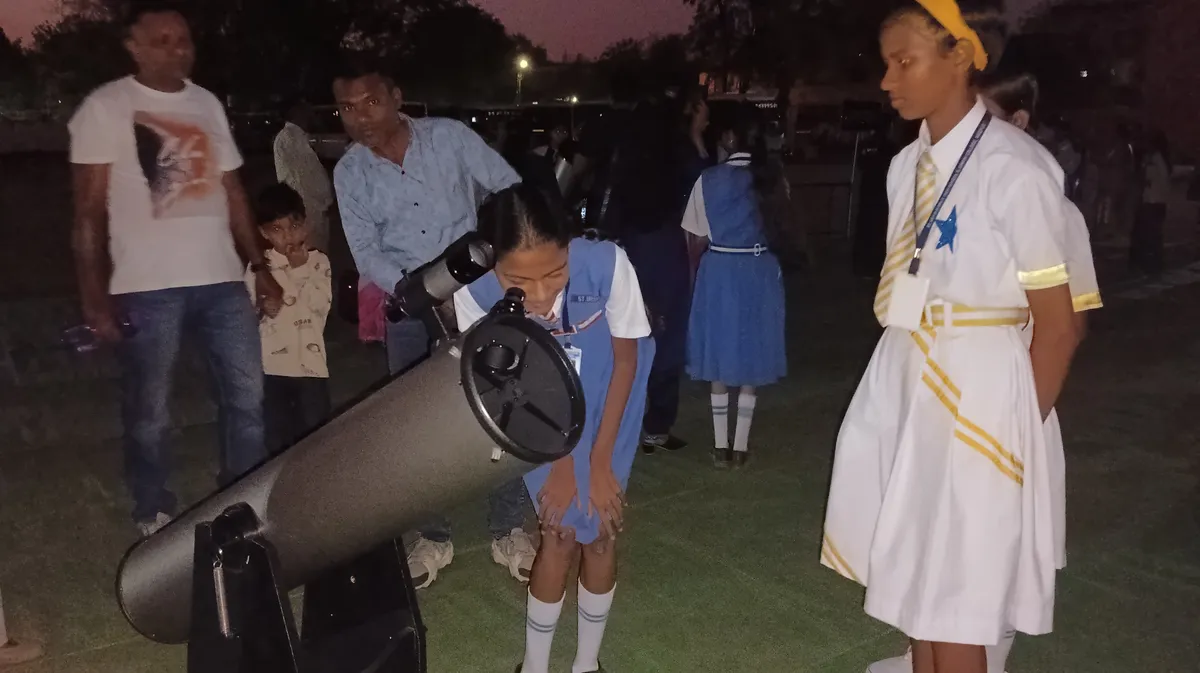 Girl in blue uniform looks through telescope while adults and children stand nearby during St. Ursula Girls' High School and Junior College sky observation programme