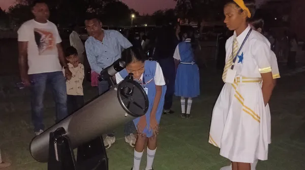 Girl in blue uniform looks through telescope while adults and children stand nearby during St. Ursula Girls' High School and Junior College sky observation programme
