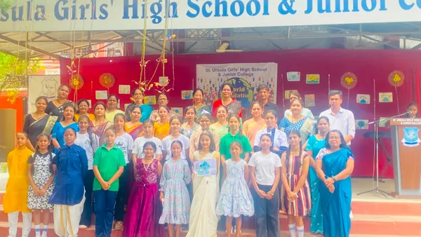 Group photo of students and teachers posing on a school stage in front of a red backdrop with a banner and a podium at St. Ursula