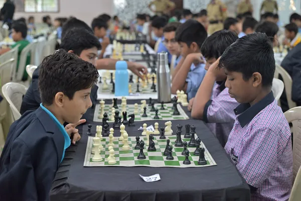 Boys playing chess at a table in a crowded hall, with other players and a few uniformed officials in the background at St. Vincent Pallotti.