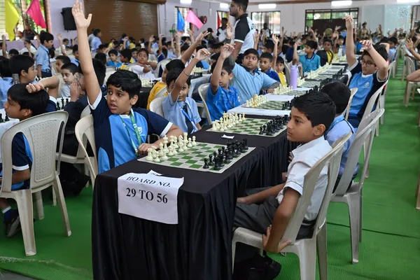 Many students in a hall at chess tables, several raising their hands, with a board number sign in front at St. Vincent Pallotti.