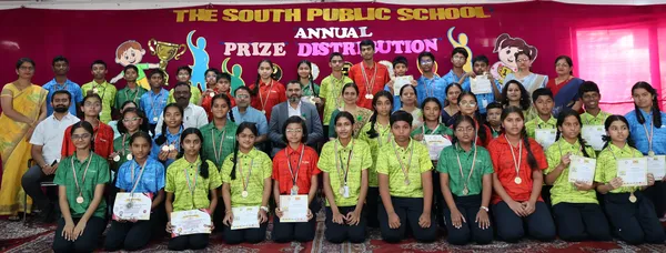 Group photo of students wearing medals and holding certificates posing with adults in front of a prize distribution stage backdrop, The South Public School.